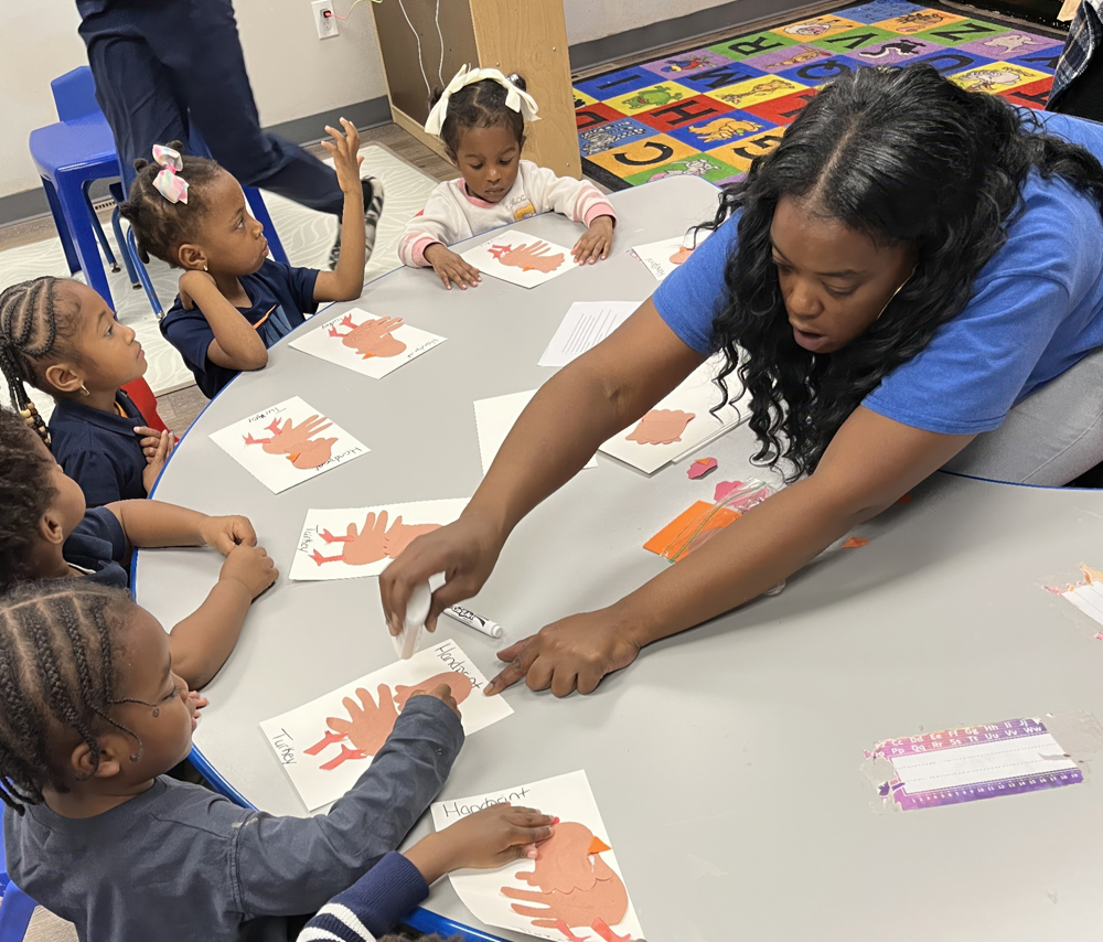 A teacher guides children in a creative activity, helping them with handprint crafts at a table.