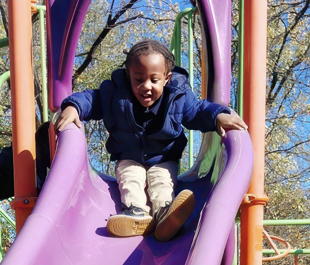 A young child joyfully slides down a slide at a playground, wearing a blue jacket and smiling as they enjoy the moment.