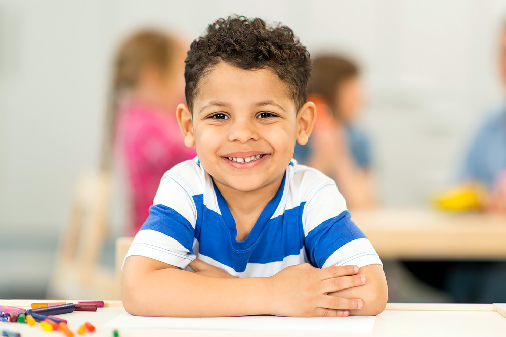 A smiling young boy with curly hair leans on a table in a classroom, radiating joy and confidence.