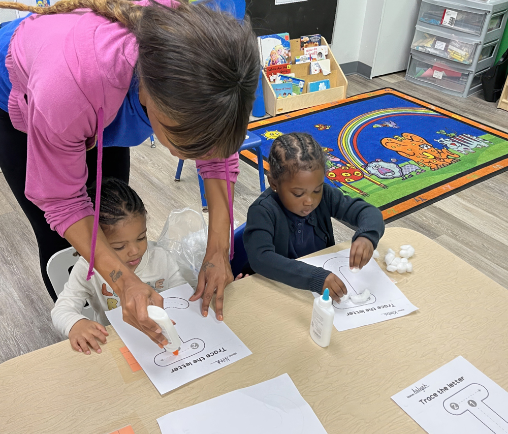 A teacher assists two children as they craft with glue and cotton balls, focusing on their creative activity at a table.