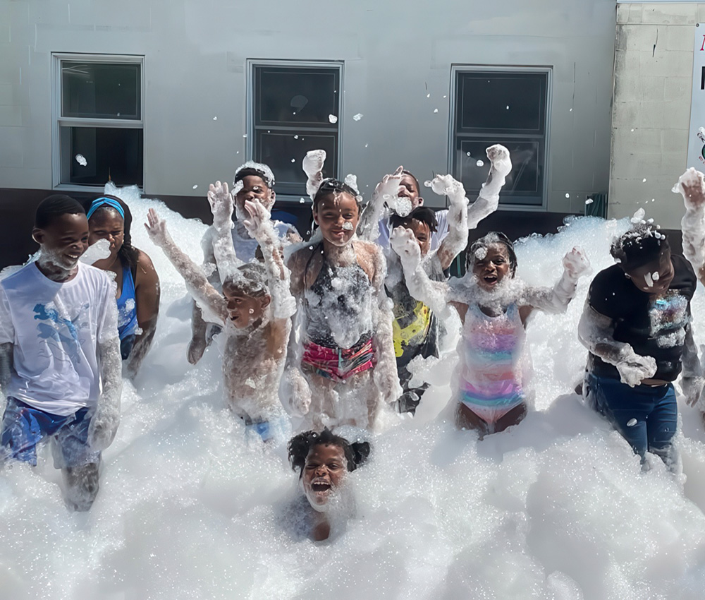 Kids joyfully play in a sea of bubbles, laughing and splashing in a fun, frothy outdoor setting.