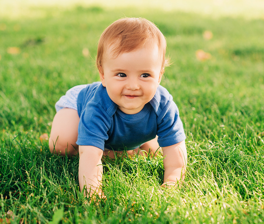 A playful baby crawls on the grass, grinning with delight, enjoying a sunny day outdoors.