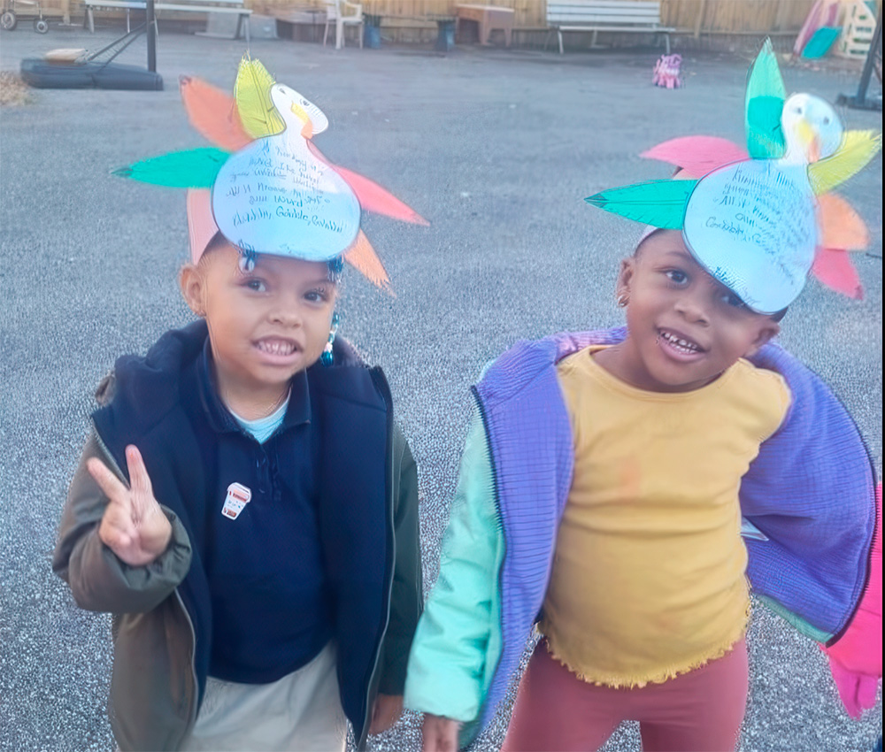 Two smiling children wear colorful paper hats adorned with feathers, posing playfully outdoors while showing peace signs.