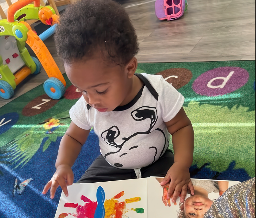 A focused toddler in a Snoopy t-shirt explores a colorful book, sitting on a playful rug, engaged in learning and discovery.