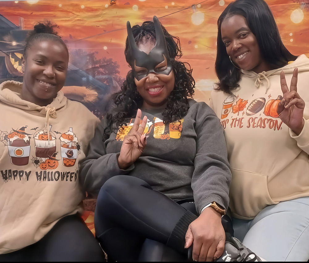 Three teacher pose playfully, one wearing a bat-themed mask, all in cozy hoodies, against a colorful autumn backdrop.