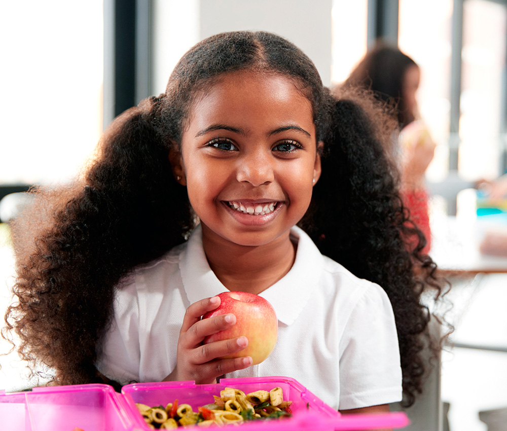 A girl with curly hair smiles brightly while holding an apple, enjoying a healthy snack in a cheerful setting.