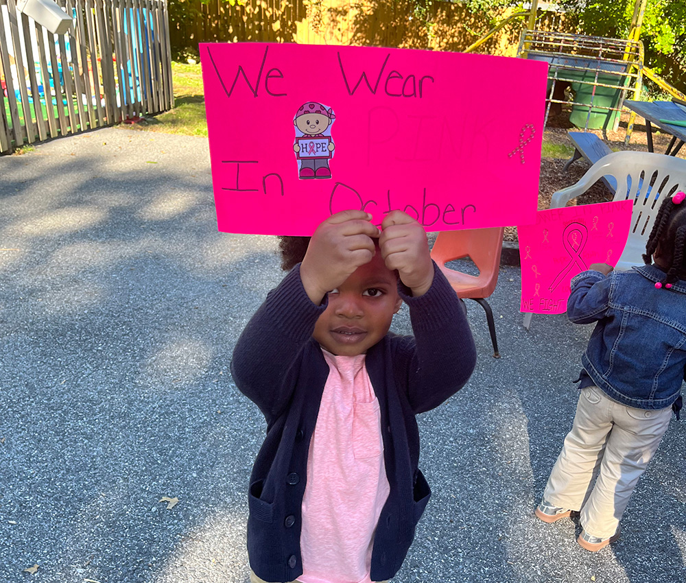 A child proudly holds up a bright pink sign that reads "We Wear Pink in October," participating in a meaningful event.