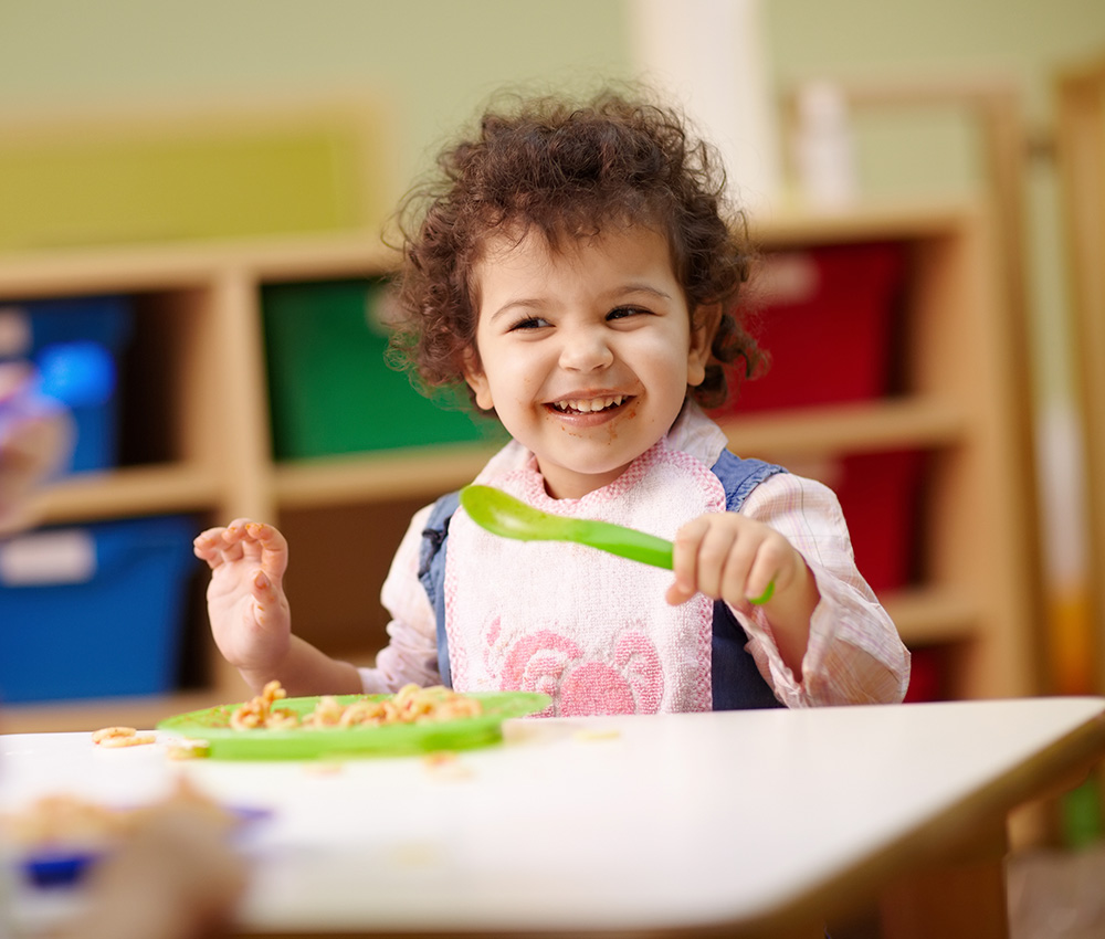 A cheerful toddler smiles while eating, holding a green spoon, in a colorful and playful dining environment.