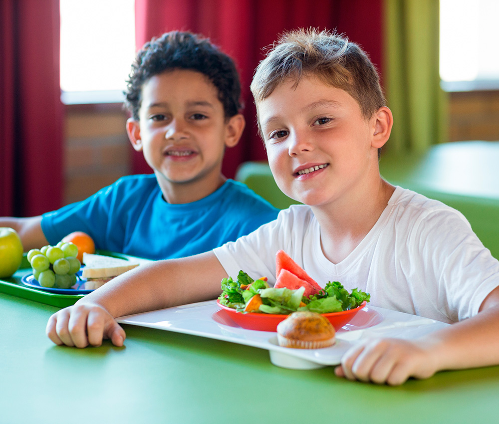 Two boys smile with healthy meals in front of them, enjoying a fun dining experience together.