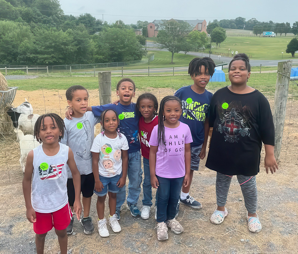 A group of kids smiles together at a farm, wearing stickers and enjoying a fun day outdoors with animals.