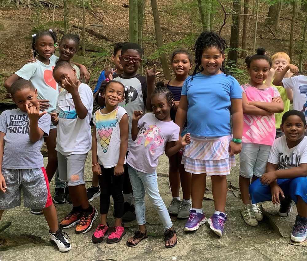 A group of children poses together in a forest, smiling and showing playful gestures, enjoying a fun outdoor adventure.