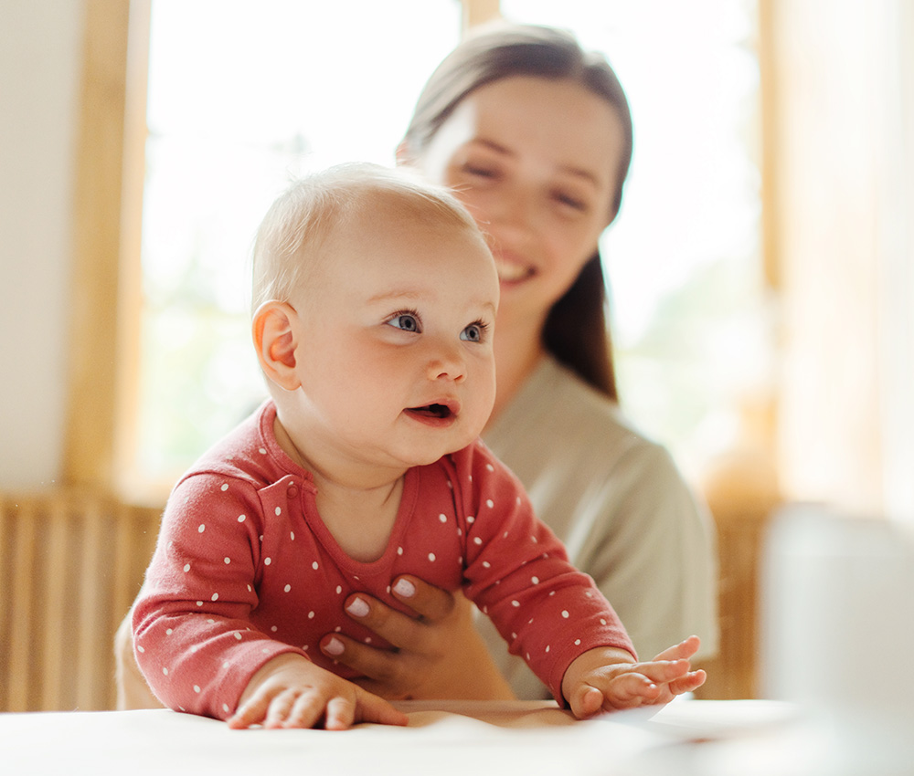 A curious baby in a polka dot outfit looks up with wide eyes, while teacher carry her, creating a warm moment.