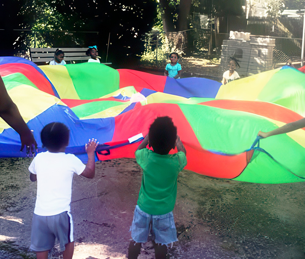 Children gather around a colorful parachute, lifting and playing with it joyfully in an outdoor setting.