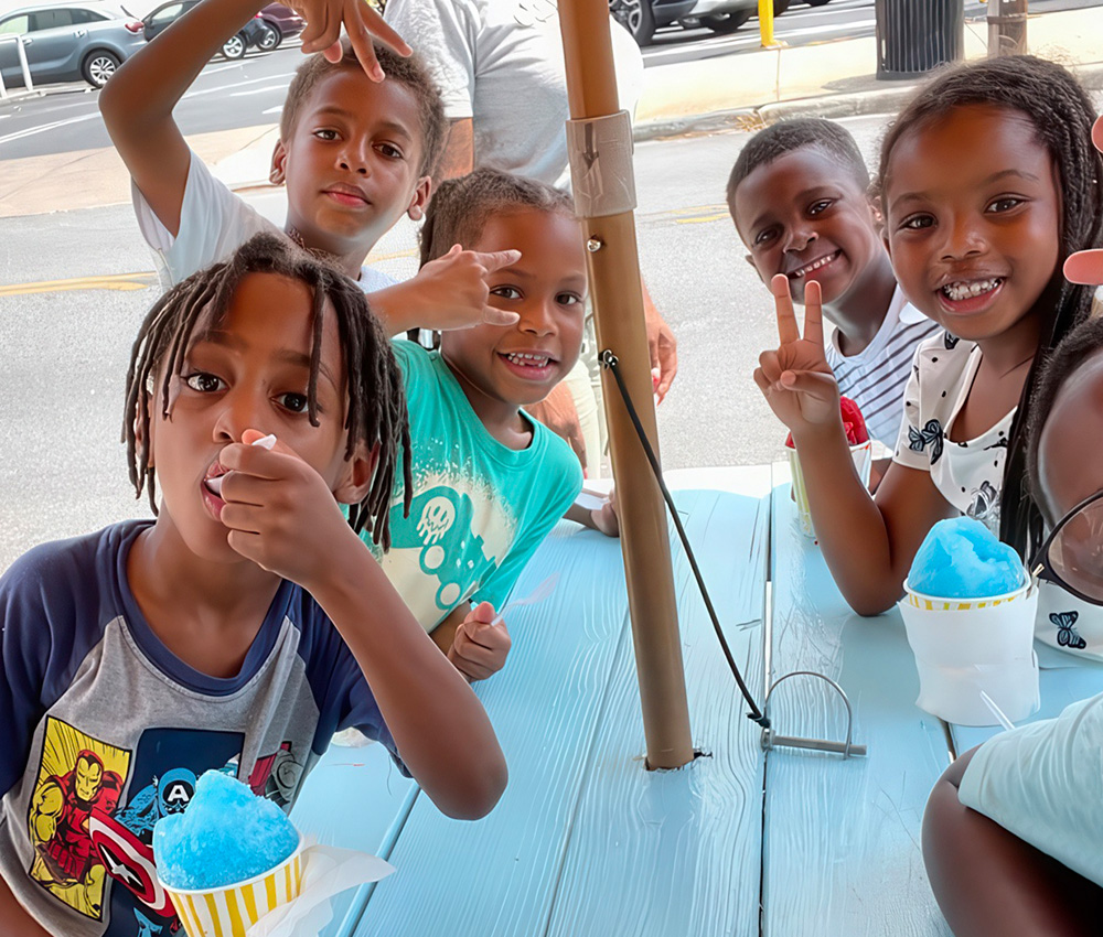 Kids gather around a table, happily enjoying snow cones and posing with cheerful smiles and playful gestures.