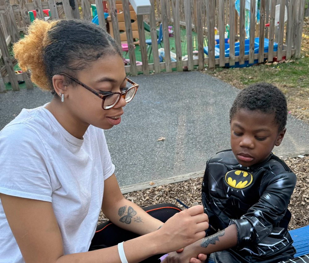 A teacher helps a boy dressed in a Batman costume, engaged in a caring moment outdoors in a playful setting.