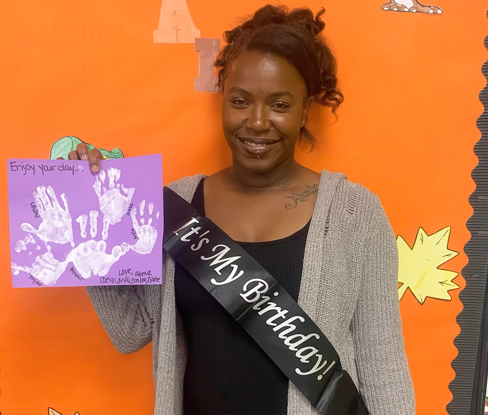 A teacher wearing a "It's My Birthday!" sash smiles while holding a colorful birthday card, set against a festive backdrop.