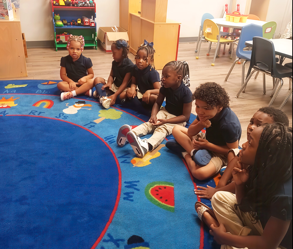 A group of young children sits in a circle on a colorful carpet, attentively participating in a classroom activity.