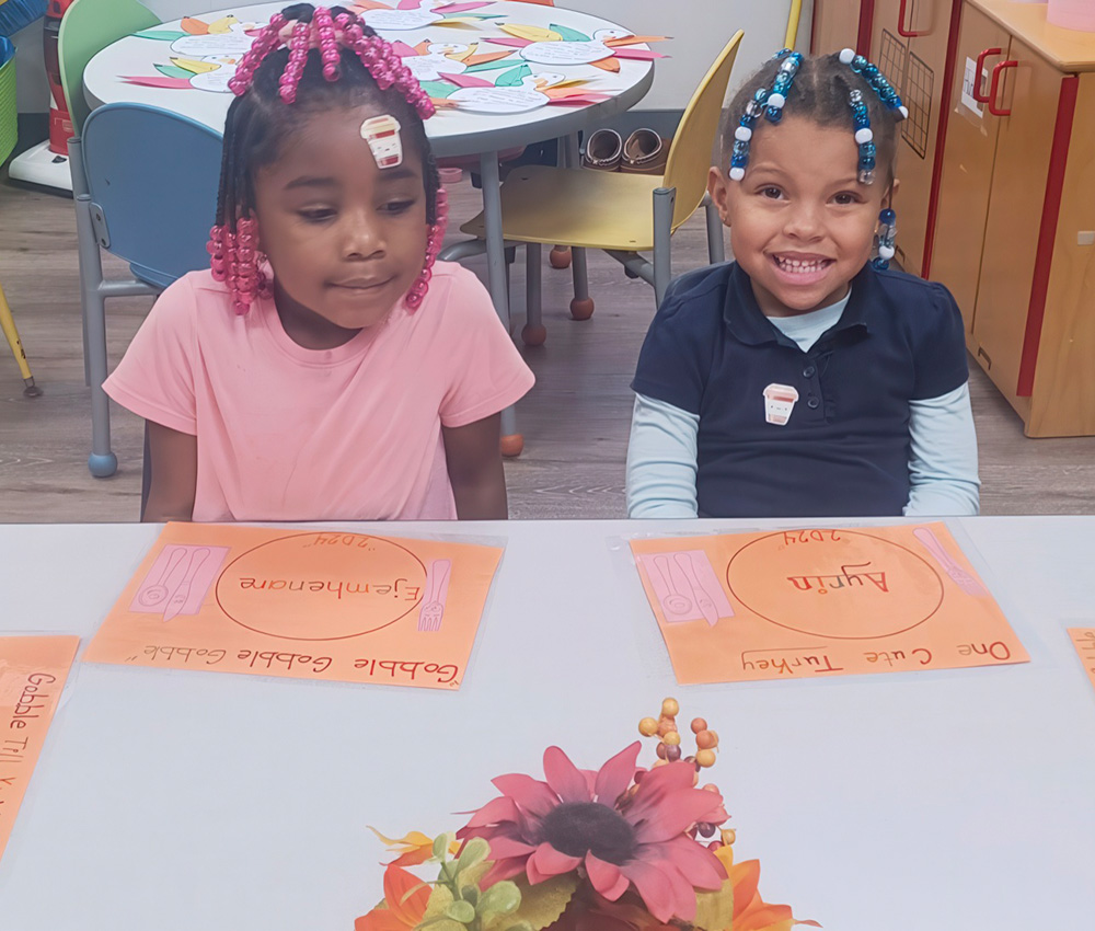 Two girls sit at a table, each with a colorful placemat, smiling and ready for an activity in a cheerful classroom.