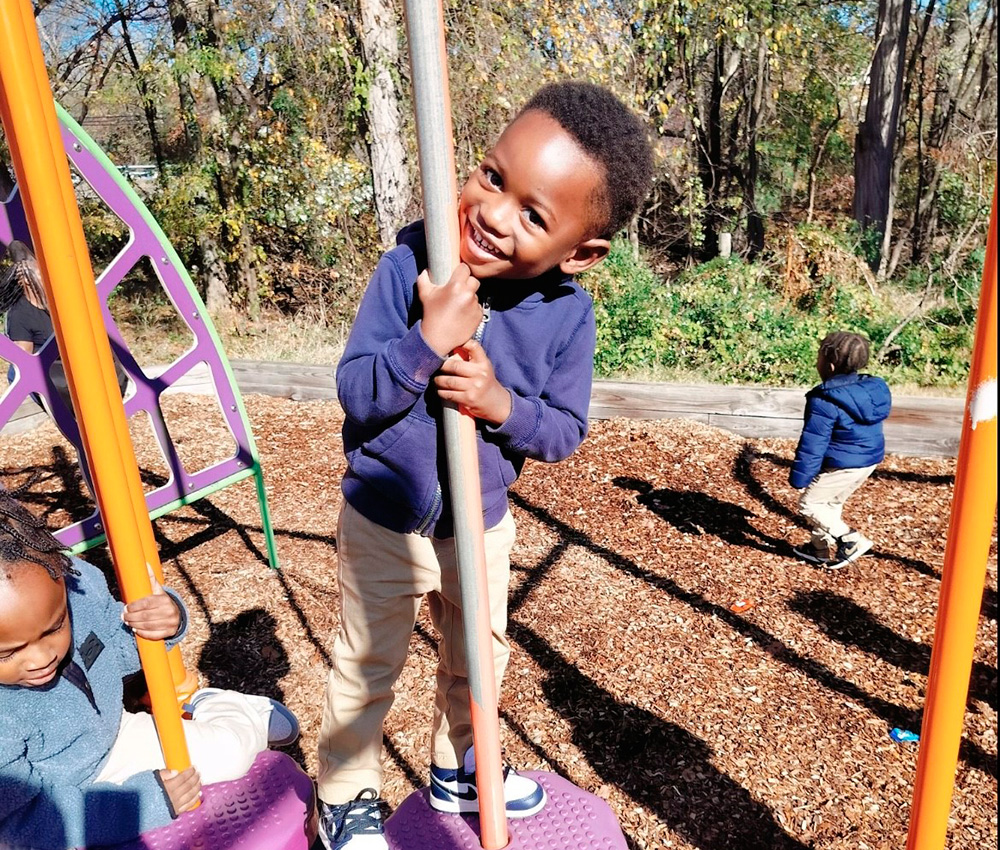 A cheerful young boy smiles while holding onto a playground pole, enjoying playtime in a sunny outdoor environment.