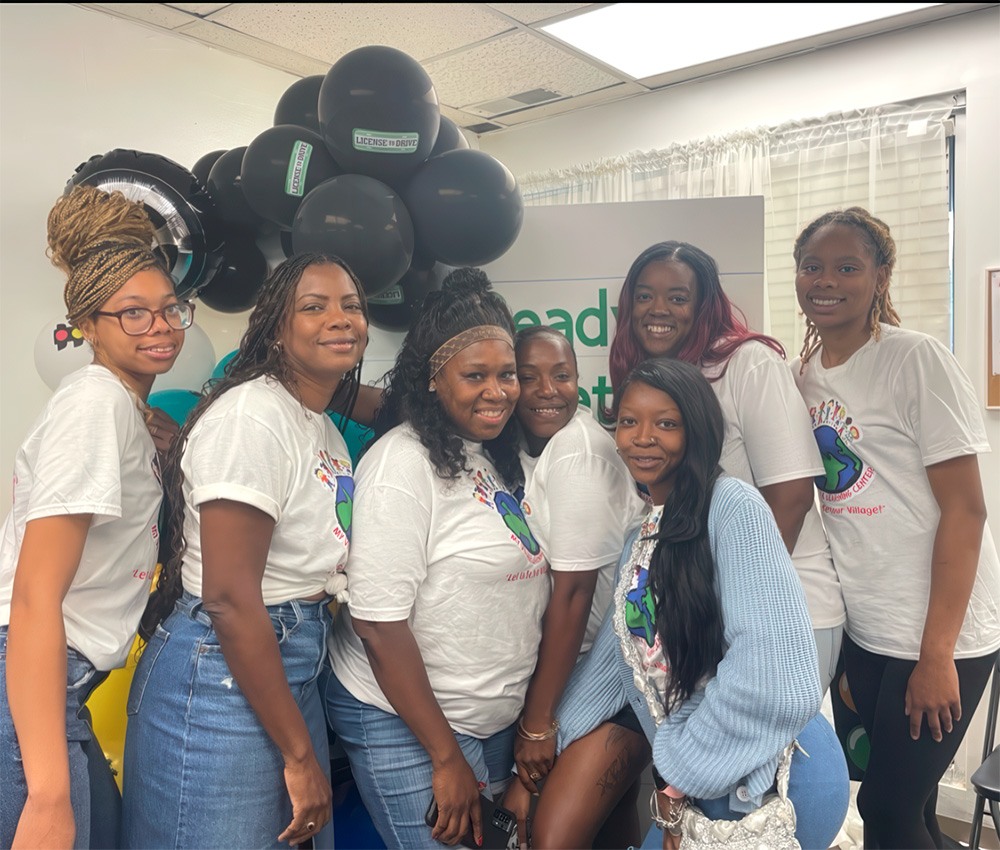 A group of smiling teachers stands together, wearing matching shirts, in front of black balloons and a cheerful backdrop.