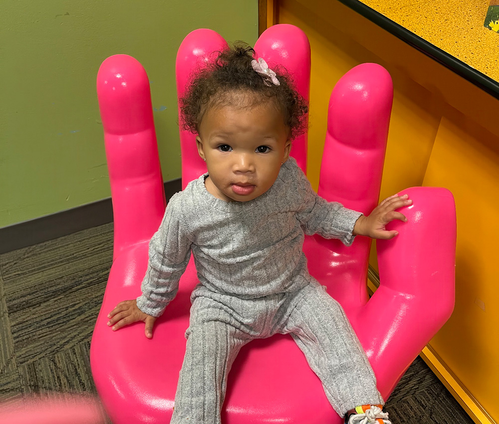 A cute toddler sits on a giant pink hand sculpture, looking curiously at the camera in a playful setting.