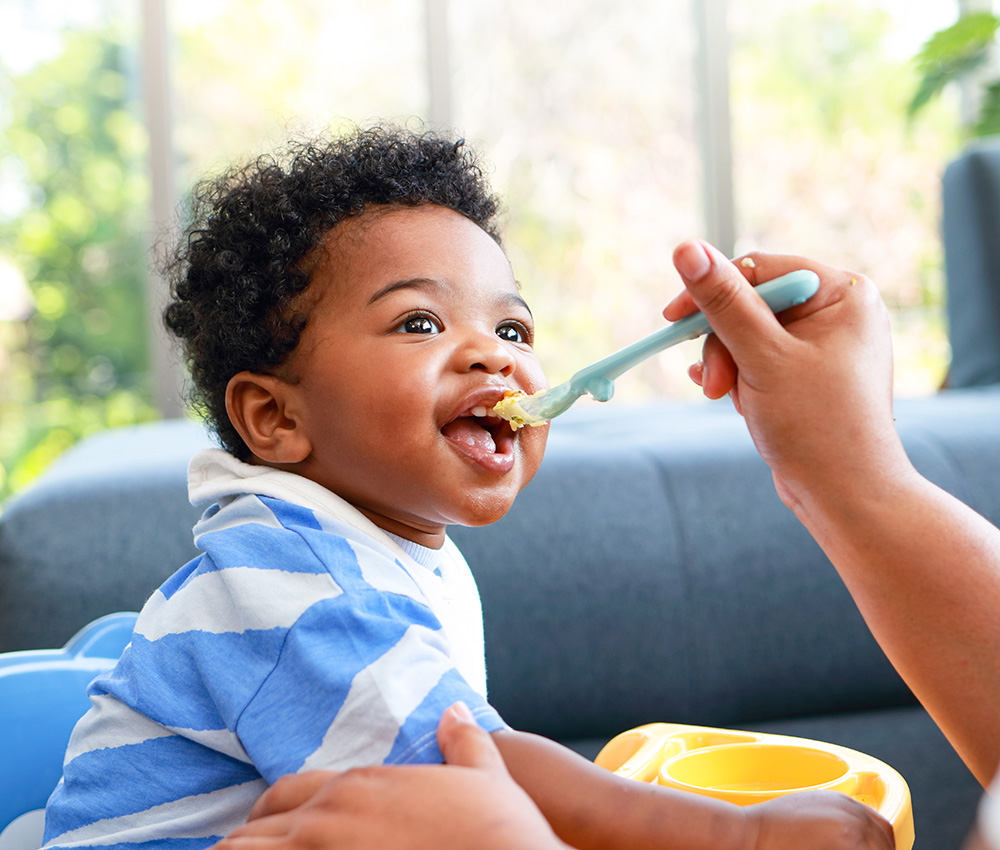 A happy toddler in a striped shirt eagerly opens their mouth for a spoonful of food, enjoying mealtime with a caring adult.