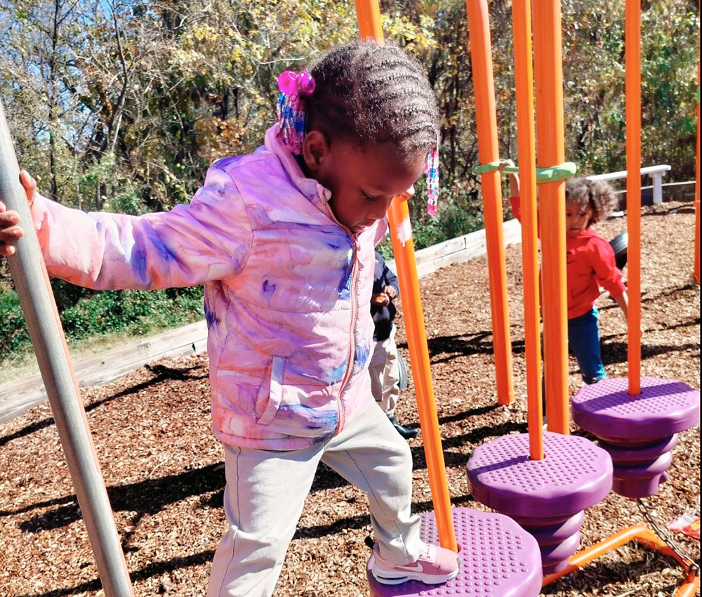 A girl explores a playground, balancing on equipment with a look of focus and determination in a sunny outdoor setting.