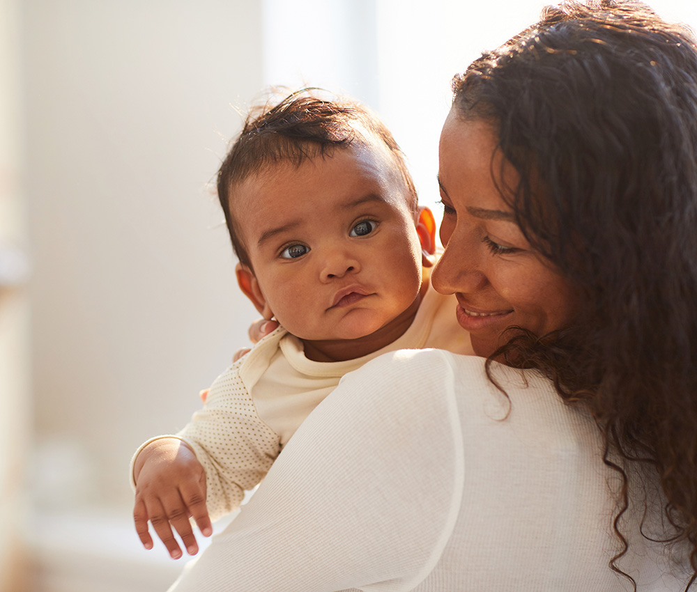 A tender moment as a mother holds her baby close, both sharing a calm, loving gaze in a softly lit room.