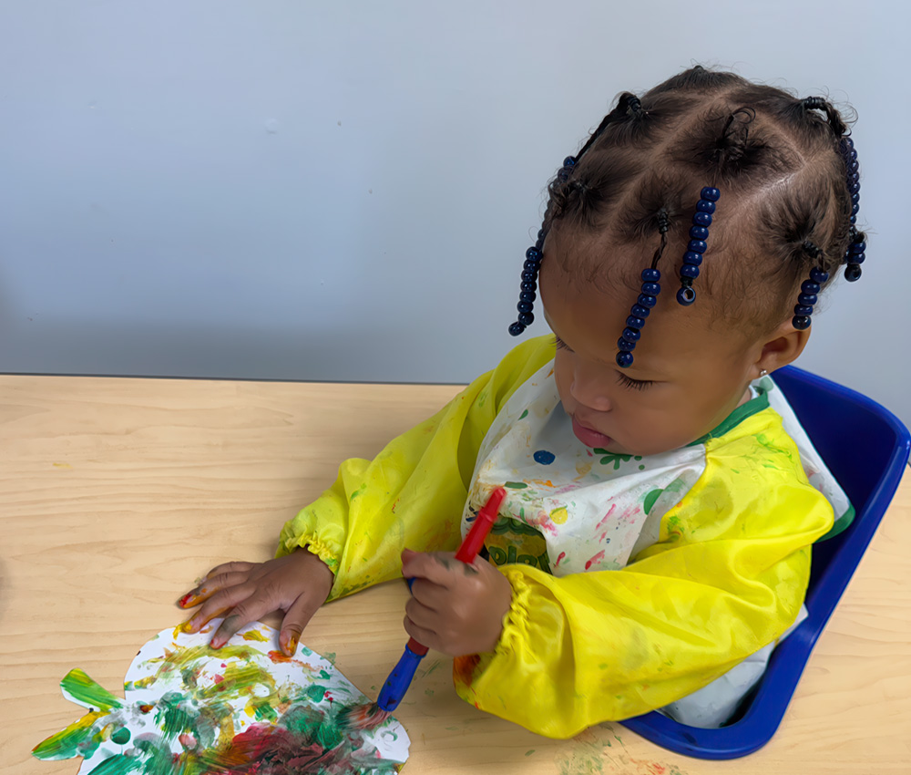 A focused toddler in a colorful apron paints with a brush, immersed in creativity at a bright and fun art station.