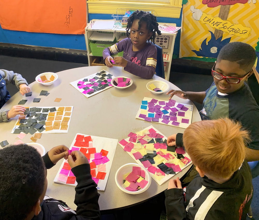 Children work together on a craft, arranging colorful paper pieces into patterns at a circular table.