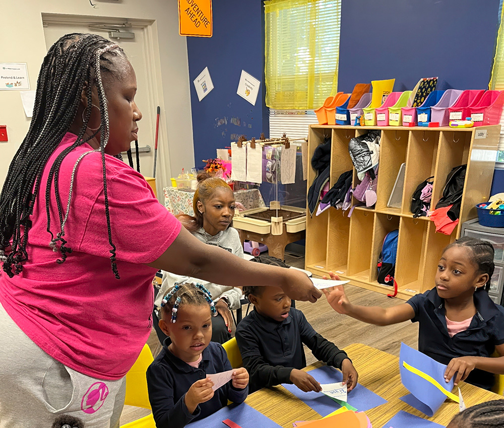 A teacher hands out materials in a classroom, while younger students eagerly reach out to receive them showing collaboration.