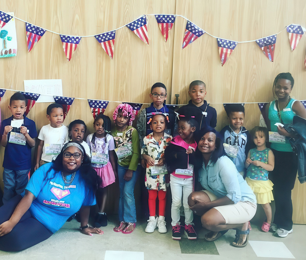 A joyful group of children and teachers smiling in front of festive American flags, celebrating community and togetherness.