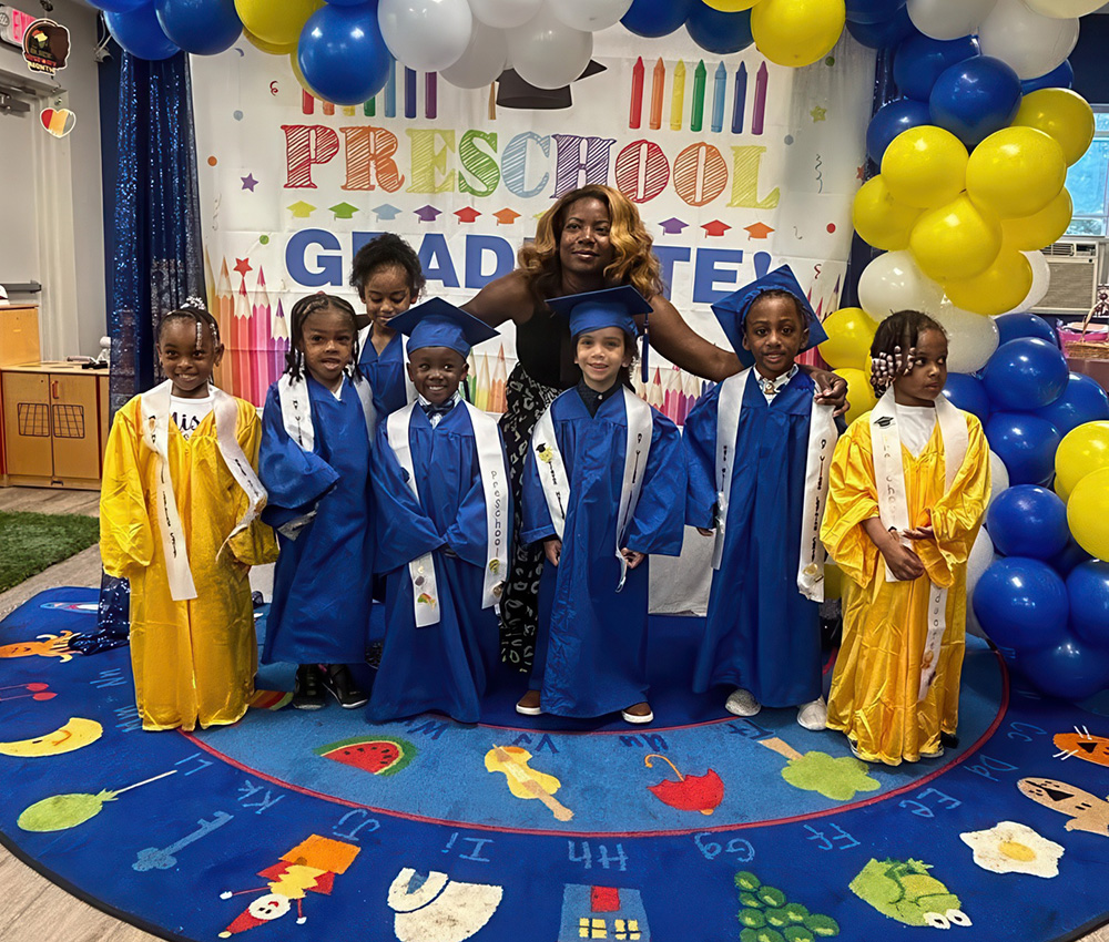 Kids in graduation gowns celebrate preschool graduation with their teacher, smiling in front of colorful decorations.