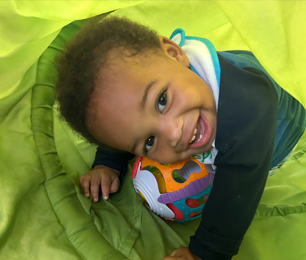 A happy toddler peeks out while playing with a colorful ball inside a green play tent, radiating joy and curiosity.