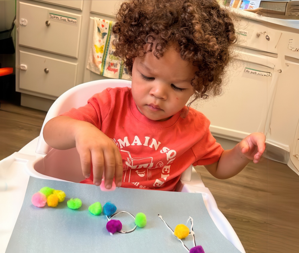 A young child with curly hair carefully places colorful pom-poms onto a craft project, focused and engaged in creative play.