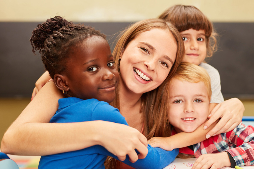 A group of children and a teacher share a warm embrace, all smiling happily together in a cozy indoor setting.