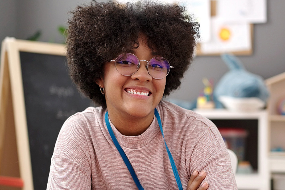 A smiling young teacher with curly hair and glasses poses confidently, showcasing a friendly demeanor in a classroom setting.