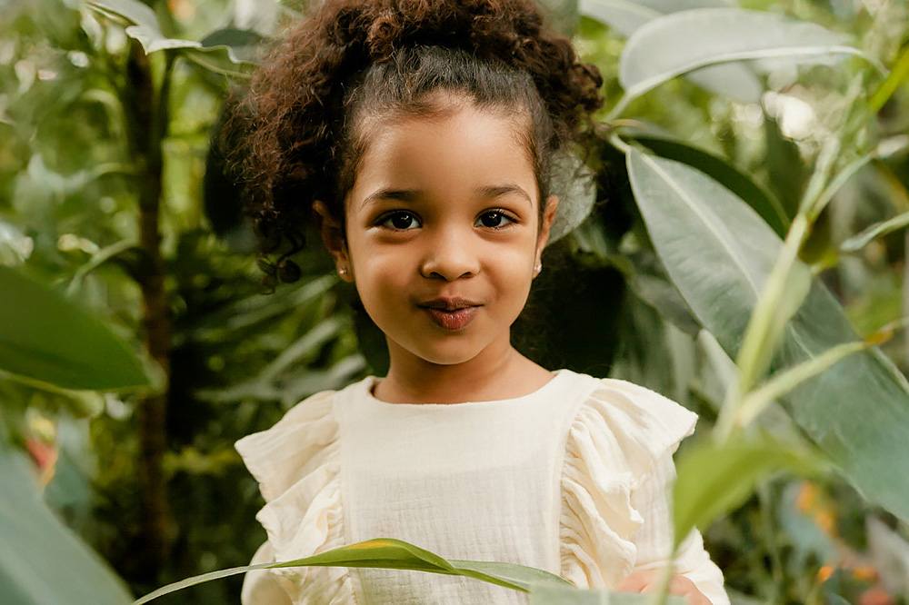 A young girl with curly hair playfully peeks through lush green leaves, exuding a sense of wonder in a natural setting.