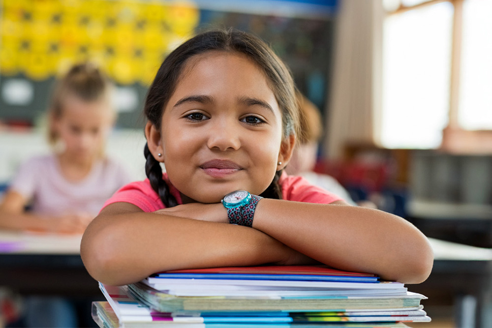 A girl with braids smiles confidently while resting her arms on a stack of books in a classroom setting.