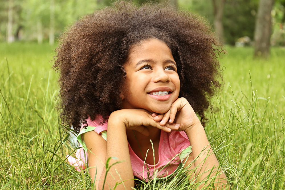 A girl lies in the grass, smiling and daydreaming, surrounded by nature's beauty on a sunny day.