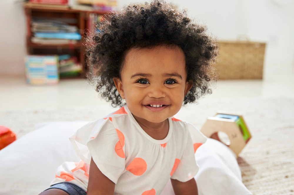 A smiling toddler with curly hair sits on the floor, wearing a polka dot shirt, radiating joy in a playful environment.