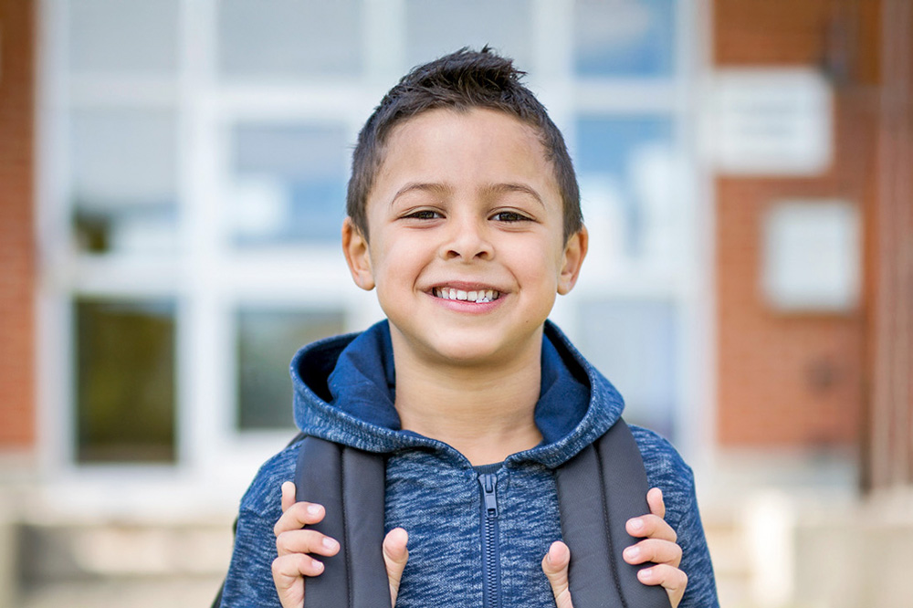 A smiling boy with a backpack stands in front of a school, ready for the day.