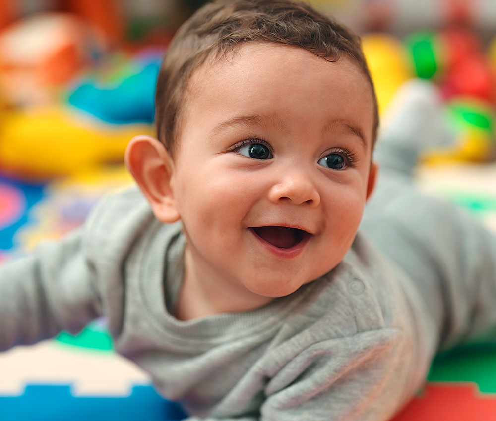 A joyful baby in a gray outfit smiles widely while playing on a colorful mat, radiating happiness.