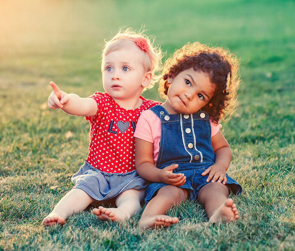 Two toddlers sit together on the grass, one pointing excitedly while the other looks on with a curious expression.