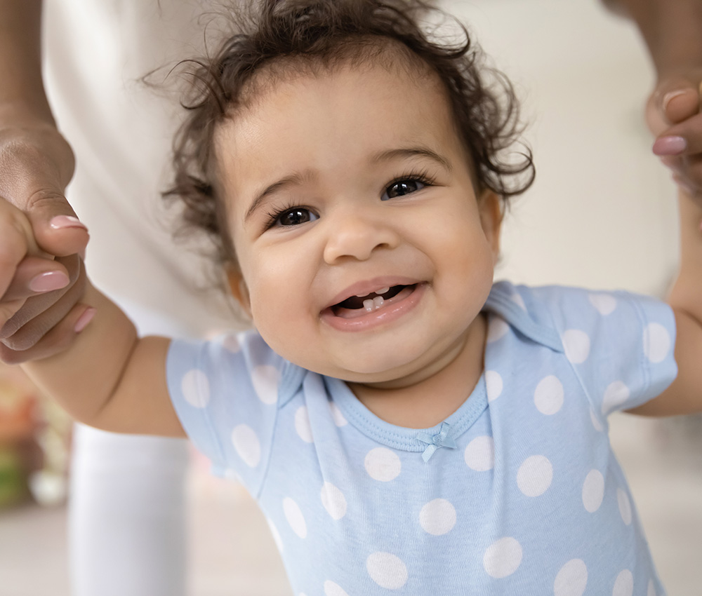 A joyful baby in a polka dot onesie beams while being held up, showcasing a big smile and a sense of excitement.