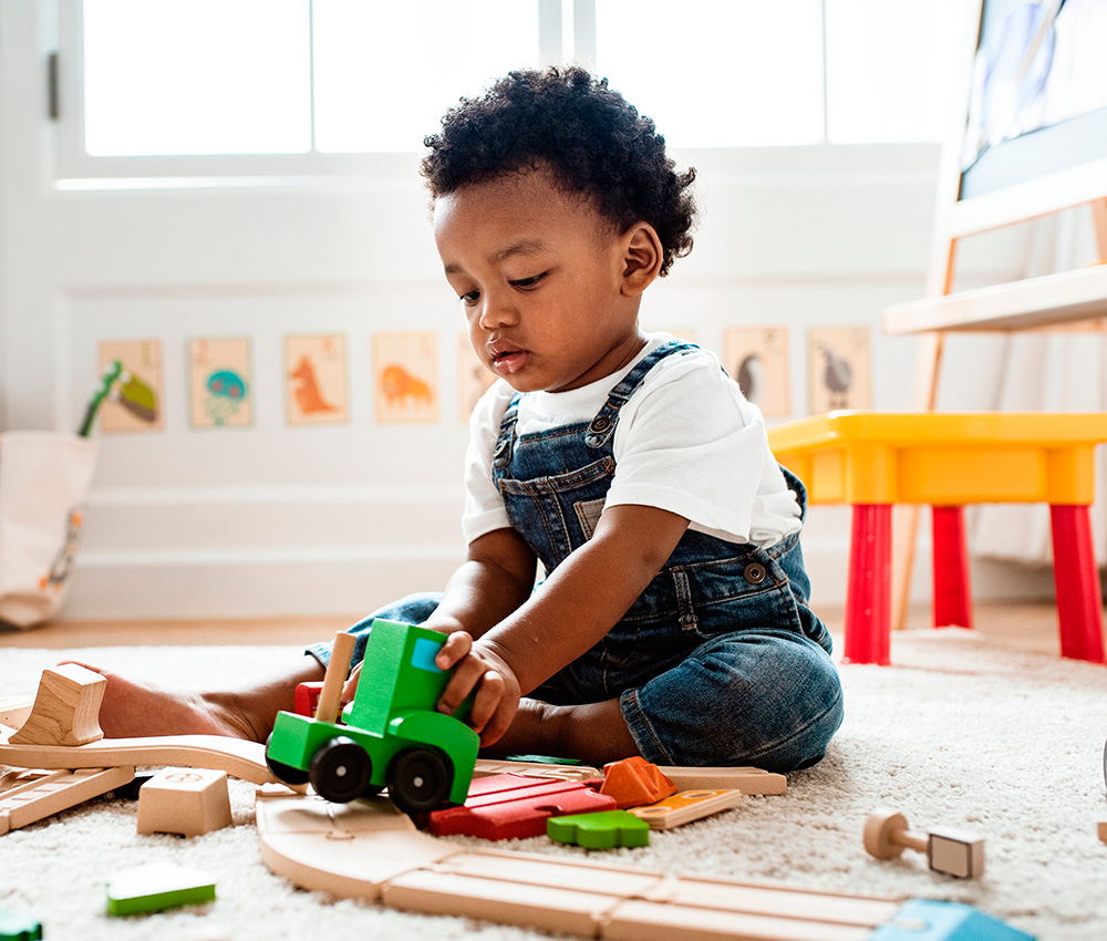 A toddler in overalls plays with colorful wooden toys, deeply engaged in imaginative play in a bright room.