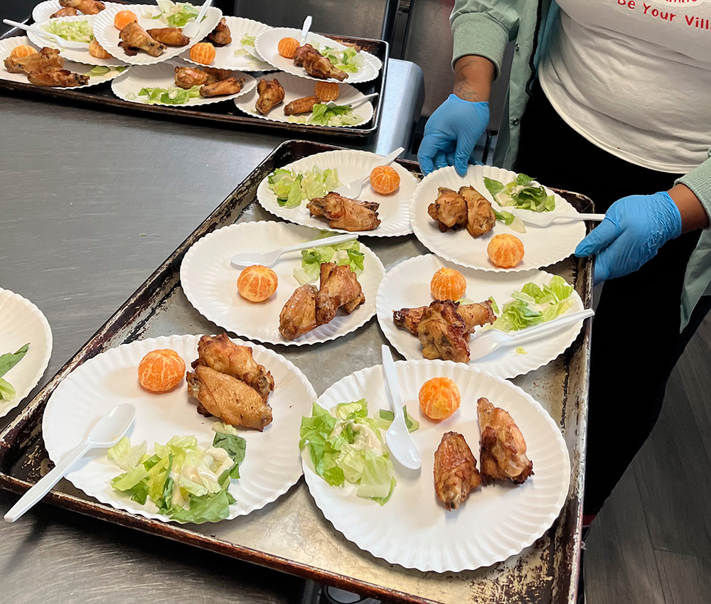 Plates of food are being prepared, featuring chicken wings, lettuce, and orange slices, arranged neatly on paper plates.