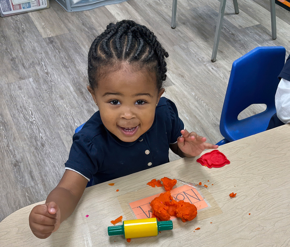 A joyful toddler smiles while playing with orange playdough at a table, surrounded by colorful tools and creative materials.