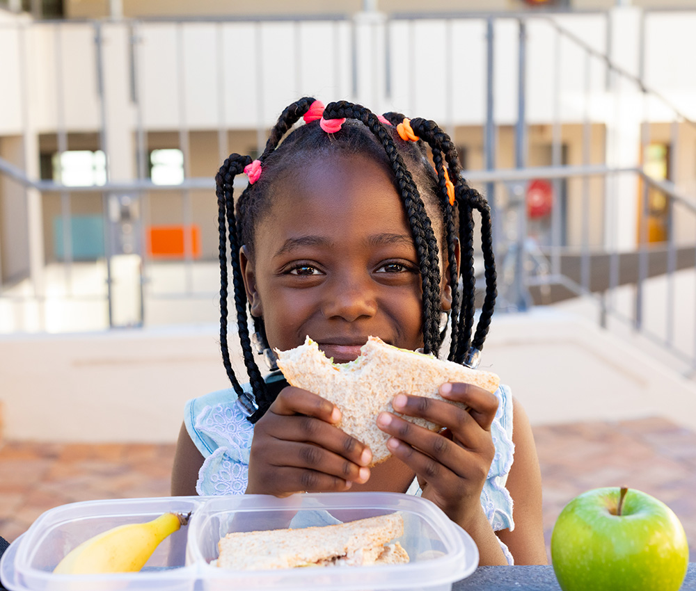 A girl smiles brightly while holding a sandwich, enjoying a delightful snack during a sunny day outdoors.